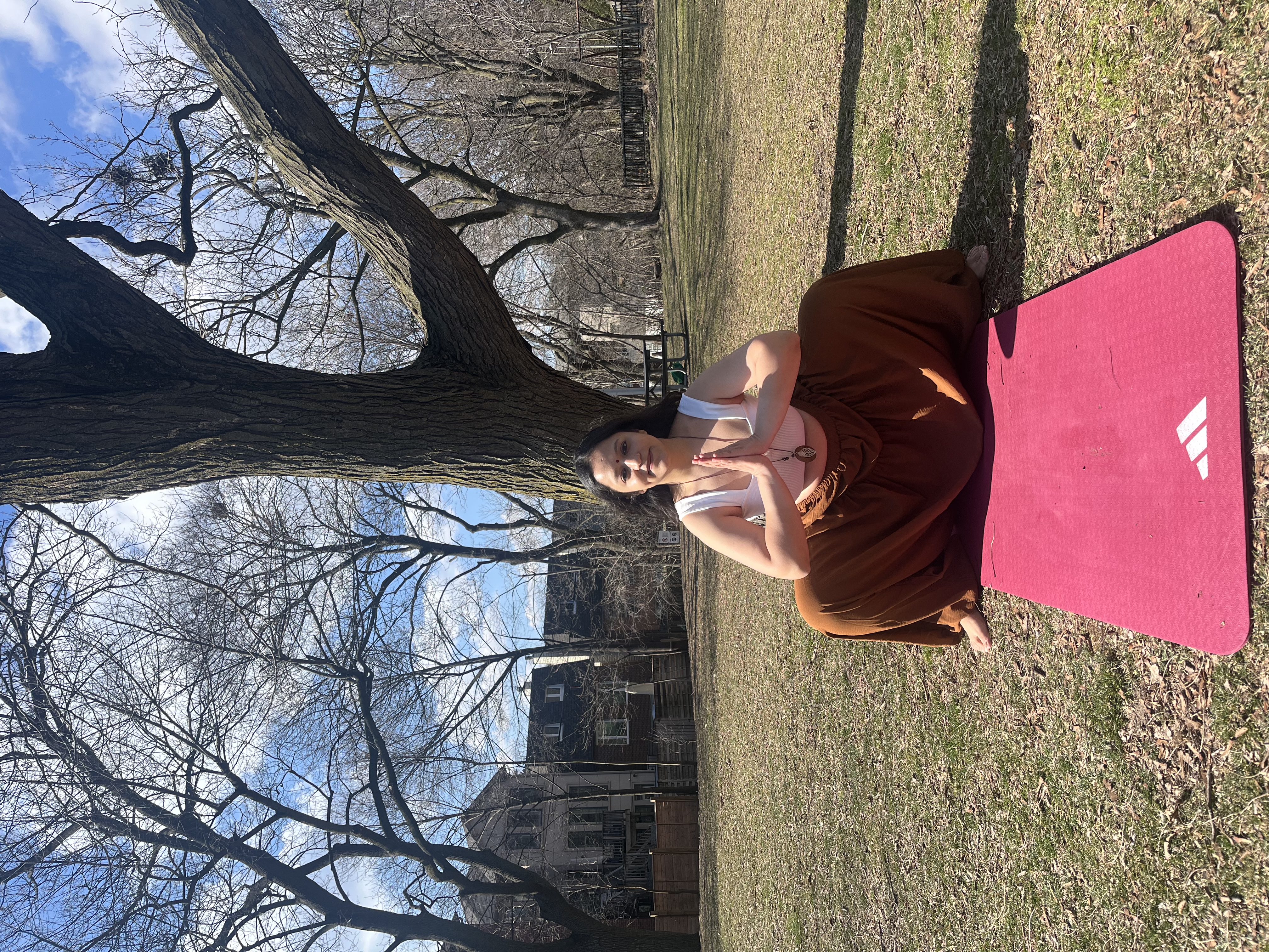 Yashna seated in namaste on the grass beneath a large tree