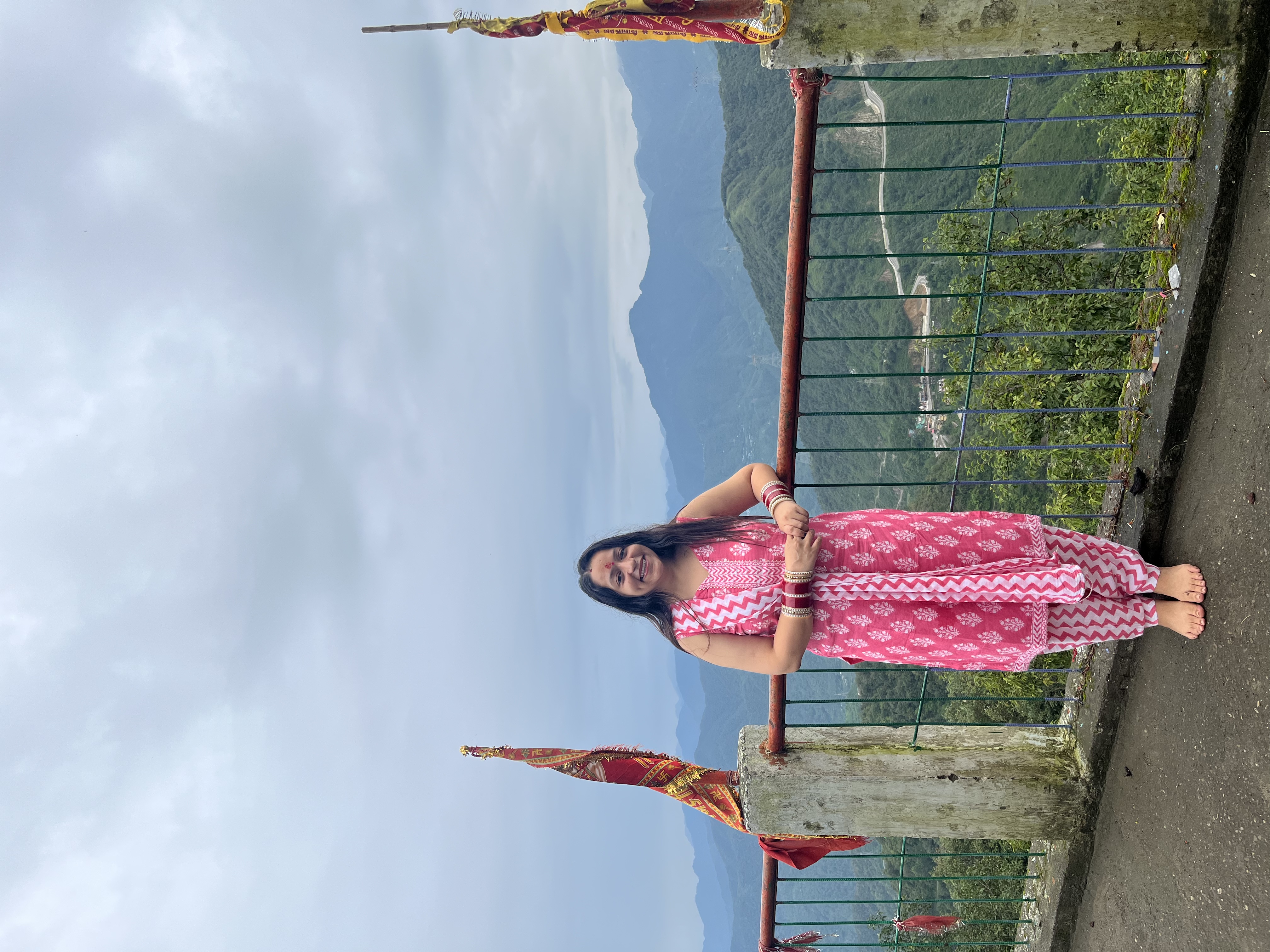 Yashna at a mountain overlook, standing by a railing under open sky